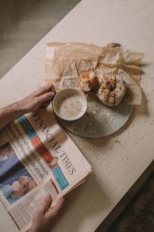 A person holds a newspaper titled 'Financial Times' with headlines and images related to current events. Beside the newspaper, there is a cup of coffee with foam on top and a plate containing two pastries. The table surface is light-colored and has crumbs scattered on it.