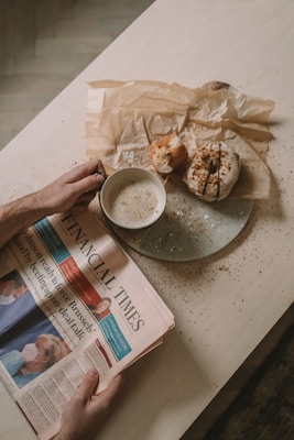 A person holds a newspaper titled 'Financial Times' with headlines and images related to current events. Beside the newspaper, there is a cup of coffee with foam on top and a plate containing two pastries. The table surface is light-colored and has crumbs scattered on it.