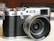A vintage-style digital camera with a textured black grip and silver metallic accents, featuring a prominent lens with visible markings. The camera is placed on a light-colored surface, with blurred background elements including a stack of papers.