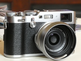 A vintage-style digital camera with a textured black grip and silver metallic accents, featuring a prominent lens with visible markings. The camera is placed on a light-colored surface, with blurred background elements including a stack of papers.