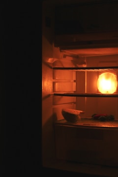 A dimly lit refrigerator with a glowing light on the inside. On a shelf, there is a piece of bread or food item partially unwrapped, with some greens next to it. The overall atmosphere is dark with warmth emanating from the light bulb inside.
