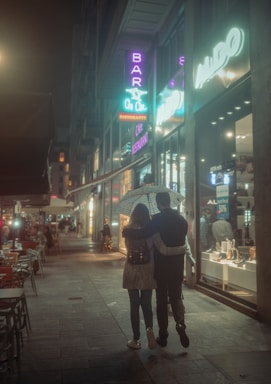 A couple walks under an umbrella along a dimly lit city street at night. Neon signs illuminate the scene with soft purple and blue hues, and shop windows display various items. The atmosphere is cozy and intimate, with outdoor tables lining the sidewalk.