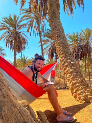 A man is sitting in a colorful hammock under tall palm trees. He is smiling and holding a laptop, wearing flip-flops on a sandy ground. The sky is clear and blue, suggesting a sunny day.