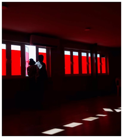 Two individuals stand silhouetted against a series of large windows with bright red panels. The light casts striking reflections on the floor, creating patterns in the dimly lit room. The scene suggests introspection or contemplation.