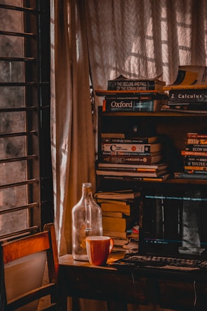 A cozy study corner with a wooden chair and a desk cluttered with books. A laptop displays a paused video scene on its screen. An empty glass bottle and a striped mug are placed on the desk. Natural light filters through curtains, giving a warm, inviting ambiance.