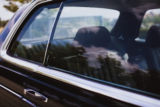 A close-up view of a car window reflecting the surrounding trees and sky. The interior of the car is visible, showcasing leather seats with no occupant inside. The window allows for a partial view of the interior while the reflections create an overlay effect.