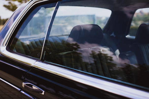 A close-up view of a car window reflecting the surrounding trees and sky. The interior of the car is visible, showcasing leather seats with no occupant inside. The window allows for a partial view of the interior while the reflections create an overlay effect.