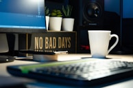 A workspace featuring a computer monitor displaying code, a white coffee mug, a smartphone, and a sign with the message 'NO BAD DAYS'. Potted plants and a speaker add to the decor, creating a calming and motivational atmosphere.