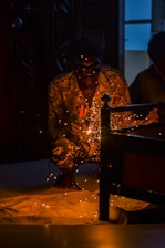 A person is crouched down wearing goggles, engaged in welding with sparks flying around. The setting is dimly lit, focusing on the glowing sparks and the welding activity. The figure is wearing a patterned shirt and is partially obscured by shadows, with some background details of furniture and a door visible.