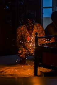 A person is crouched down wearing goggles, engaged in welding with sparks flying around. The setting is dimly lit, focusing on the glowing sparks and the welding activity. The figure is wearing a patterned shirt and is partially obscured by shadows, with some background details of furniture and a door visible.