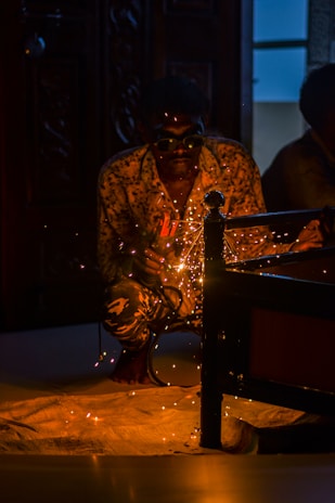 A person is crouched down wearing goggles, engaged in welding with sparks flying around. The setting is dimly lit, focusing on the glowing sparks and the welding activity. The figure is wearing a patterned shirt and is partially obscured by shadows, with some background details of furniture and a door visible.
