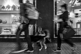 A homeless person sits against a wall, holding a sign and accompanied by a dog. Blurred figures of pedestrians pass by quickly, highlighting the contrast between movement and stillness. Storefront items are displayed in the window behind the person, with various decorative objects. The image is in black and white, emphasizing a somber tone.