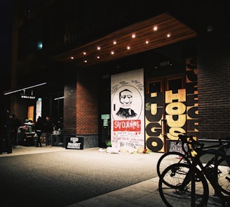 The image depicts an urban scene at night with a brick building. In front of the building, there is a large mural with a bearded man's face and the words 'SAY OUR NAMES' in bold red letters, surrounded by the names of several people. There are bicycles parked in the foreground, and a group of people standing to the left, appearing to participate in some kind of event or gathering. The building entry has prominent signage and a canopy with recessed lighting.