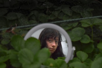 A person's face is reflected in a round mirror surrounded by lush green foliage, creating a natural and introspective scene.