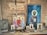A wooden shelf displays religious icons and paintings, primarily depicting figures with halos, likely saints or religious figures. A string of rosary beads and various religious artifacts are arranged on the shelf alongside the images. Above, a wooden plaque with an inscription hangs on the wall.