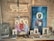 A wooden shelf displays religious icons and paintings, primarily depicting figures with halos, likely saints or religious figures. A string of rosary beads and various religious artifacts are arranged on the shelf alongside the images. Above, a wooden plaque with an inscription hangs on the wall.
