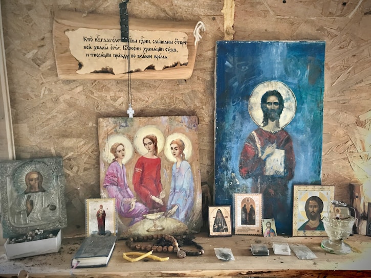 A wooden shelf displays religious icons and paintings, primarily depicting figures with halos, likely saints or religious figures. A string of rosary beads and various religious artifacts are arranged on the shelf alongside the images. Above, a wooden plaque with an inscription hangs on the wall.