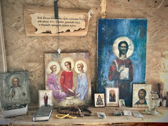 A wooden shelf displays religious icons and paintings, primarily depicting figures with halos, likely saints or religious figures. A string of rosary beads and various religious artifacts are arranged on the shelf alongside the images. Above, a wooden plaque with an inscription hangs on the wall.