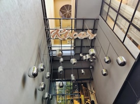 A modern architectural space with large reflective light fixtures hanging from a metal structure. The ceiling features a grid design, and a decorative sign reading 'Ricos' is attached. The space is well-lit with natural light coming through windows on the walls.