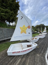A row of small white sailboats is lined up on a textured surface with green grass nearby. Each boat has a sail, with the nearest one featuring a cheerful yellow star carrying a basketball and branding for Southern Cross Travel Insurance. The sailboat is labeled 'ATLAS CONCRETE' in red letters on its side.