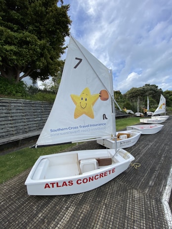 A row of small white sailboats is lined up on a textured surface with green grass nearby. Each boat has a sail, with the nearest one featuring a cheerful yellow star carrying a basketball and branding for Southern Cross Travel Insurance. The sailboat is labeled 'ATLAS CONCRETE' in red letters on its side.