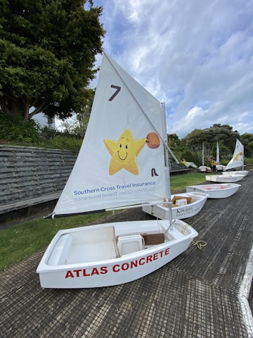 A row of small white sailboats is lined up on a textured surface with green grass nearby. Each boat has a sail, with the nearest one featuring a cheerful yellow star carrying a basketball and branding for Southern Cross Travel Insurance. The sailboat is labeled 'ATLAS CONCRETE' in red letters on its side.
