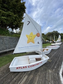A row of small white sailboats is lined up on a textured surface with green grass nearby. Each boat has a sail, with the nearest one featuring a cheerful yellow star carrying a basketball and branding for Southern Cross Travel Insurance. The sailboat is labeled 'ATLAS CONCRETE' in red letters on its side.