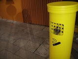 A bright yellow outdoor cigarette disposal bin is visible on a stone pavement. The bin has a black stencil of a cigarette symbol on it and several small holes on the front. A wooden paneled wall with a warm orange hue is in the background, casting shadows onto the pavement.