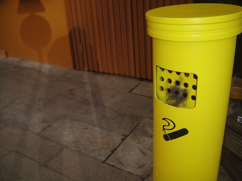 A bright yellow outdoor cigarette disposal bin is visible on a stone pavement. The bin has a black stencil of a cigarette symbol on it and several small holes on the front. A wooden paneled wall with a warm orange hue is in the background, casting shadows onto the pavement.