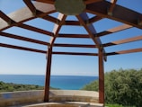 A pergola-like wooden structure frames a view of the ocean and coastline. Wooden beams form a geometric pattern against the clear blue sky. Below, a stone or concrete sitting area provides seating, with lush greenery and ocean stretching out in the background.