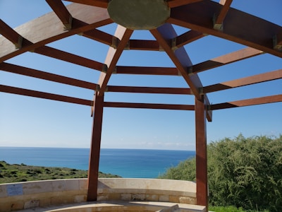 A pergola-like wooden structure frames a view of the ocean and coastline. Wooden beams form a geometric pattern against the clear blue sky. Below, a stone or concrete sitting area provides seating, with lush greenery and ocean stretching out in the background.