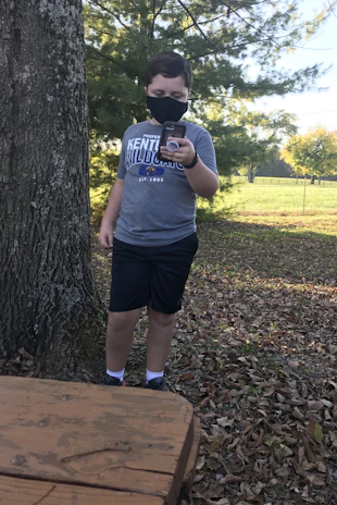 A person wearing a mask and a blue T-shirt with 'Kentucky Wildcats' printed on it is holding a smartphone near a large tree. Fallen leaves cover the ground, and there is a wooden picnic table in the foreground. The background features green trees and a fenced grassy area under a clear sky.