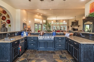 A spacious, luxurious kitchen features a large island with dark cabinetry and granite countertops. There are several pendant lights hanging from the ceiling, and the kitchen area opens into a brighter living space with windows offering a scenic view. A variety of decorative items, including a collection of colorful plates and a large statue, add character to the room.