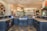 A spacious, luxurious kitchen features a large island with dark cabinetry and granite countertops. There are several pendant lights hanging from the ceiling, and the kitchen area opens into a brighter living space with windows offering a scenic view. A variety of decorative items, including a collection of colorful plates and a large statue, add character to the room.