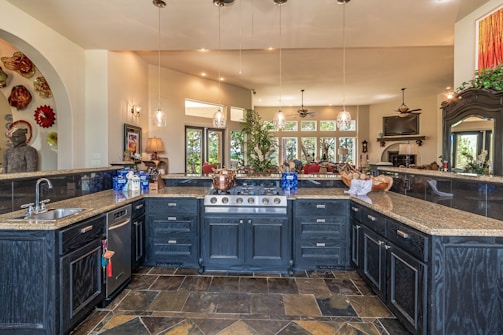 A spacious, luxurious kitchen features a large island with dark cabinetry and granite countertops. There are several pendant lights hanging from the ceiling, and the kitchen area opens into a brighter living space with windows offering a scenic view. A variety of decorative items, including a collection of colorful plates and a large statue, add character to the room.