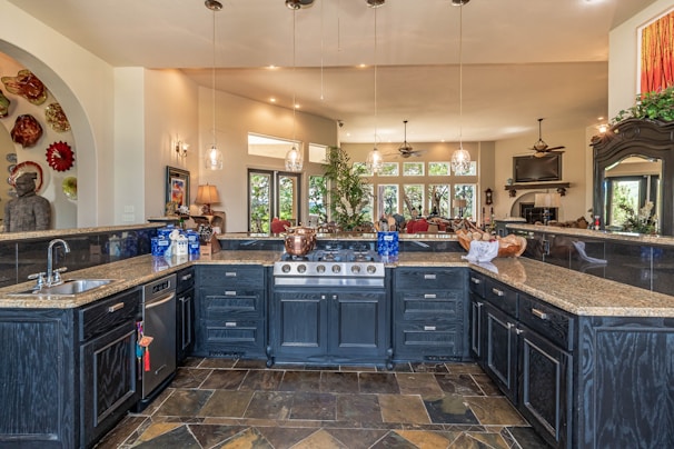 A spacious, luxurious kitchen features a large island with dark cabinetry and granite countertops. There are several pendant lights hanging from the ceiling, and the kitchen area opens into a brighter living space with windows offering a scenic view. A variety of decorative items, including a collection of colorful plates and a large statue, add character to the room.