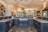 A spacious, luxurious kitchen features a large island with dark cabinetry and granite countertops. There are several pendant lights hanging from the ceiling, and the kitchen area opens into a brighter living space with windows offering a scenic view. A variety of decorative items, including a collection of colorful plates and a large statue, add character to the room.