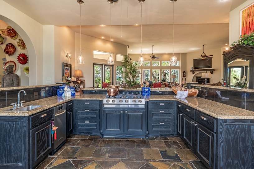 A spacious, luxurious kitchen features a large island with dark cabinetry and granite countertops. There are several pendant lights hanging from the ceiling, and the kitchen area opens into a brighter living space with windows offering a scenic view. A variety of decorative items, including a collection of colorful plates and a large statue, add character to the room.