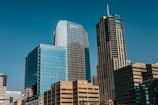 Tall modern skyscrapers with reflective glass facades against a clear blue sky, featuring multiple high-rise office buildings with visible logos such as OPTIV, POLSINELLI, and CHASE. The structures exhibit both vertical and horizontal architectural designs.