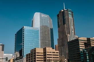 Tall modern skyscrapers with reflective glass facades against a clear blue sky, featuring multiple high-rise office buildings with visible logos such as OPTIV, POLSINELLI, and CHASE. The structures exhibit both vertical and horizontal architectural designs.
