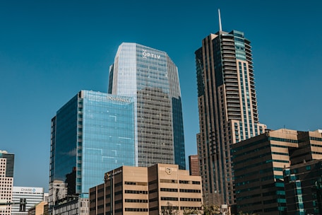 Tall modern skyscrapers with reflective glass facades against a clear blue sky, featuring multiple high-rise office buildings with visible logos such as OPTIV, POLSINELLI, and CHASE. The structures exhibit both vertical and horizontal architectural designs.