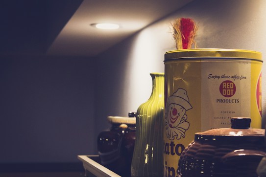 A collection of vintage containers and jars is displayed on a shelf under soft lighting. The prominent object is a yellow tin with a clown graphic and the text 'Red Dot Products.' A green vase and various brown jars accompany it, creating a nostalgic and homey atmosphere.