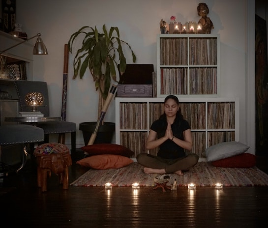 A person sits cross-legged, eyes closed, with hands in prayer position surrounded by pillows and candles. The room features shelves filled with vinyl records, a large plant, and subtle lighting from a lamp and candles. The overall setting seems cozy and serene, creating a peaceful ambiance.