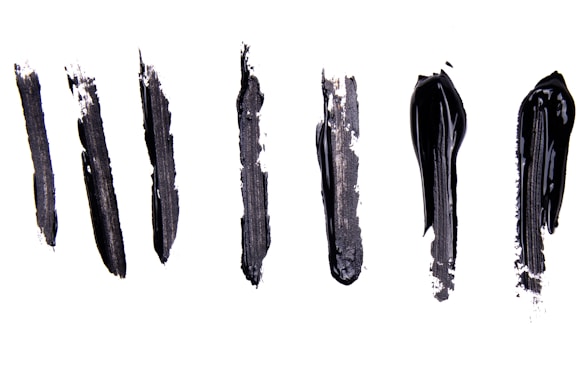 Six black brushstrokes of varying thickness and texture are arranged vertically on a white background. The strokes vary in smoothness and opacity, with some appearing more rough and textured while others are more glossy and solid.