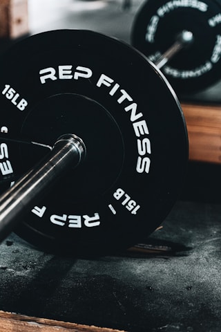 A close-up view of a barbell loaded with black weight plates labeled with 'REP FITNESS' and '15 LB'. The barbell is resting on a textured surface in a gym environment, with another barbell blurred in the background.