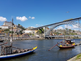 A scenic riverside view of Porto, Portugal, featuring traditional boats on the Douro River with a backdrop of colorful hillside buildings and the iconic Dom Luís I Bridge under a clear blue sky.