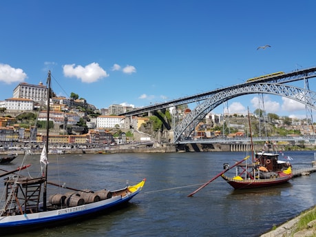 A scenic riverside view of Porto, Portugal, featuring traditional boats on the Douro River with a backdrop of colorful hillside buildings and the iconic Dom Luís I Bridge under a clear blue sky.