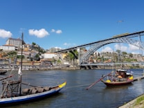 A scenic riverside view of Porto, Portugal, featuring traditional boats on the Douro River with a backdrop of colorful hillside buildings and the iconic Dom Lu&iacute;s I Bridge under a clear blue sky.