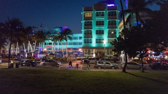 A lively nighttime scene depicts neon-lit buildings in an urban area. The building in the center features turquoise lighting, surrounded by palm trees. Cars are parked along the street, and people are strolling along the sidewalk, creating a vibrant nightlife atmosphere.