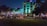 A lively nighttime scene depicts neon-lit buildings in an urban area. The building in the center features turquoise lighting, surrounded by palm trees. Cars are parked along the street, and people are strolling along the sidewalk, creating a vibrant nightlife atmosphere.